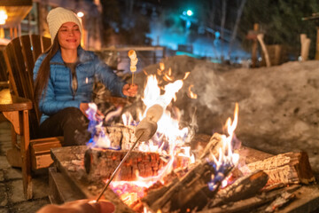 Winter campfire couple roasting marshmallows in firepit for smores at ski holiday resort. Asian happy woman relaxing after ski. - obrazy, fototapety, plakaty