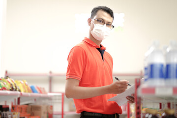  supervisor man wearing mask making notes to the clipboard while standing near a rack of mixed goods.