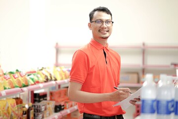 Smiling Asian young man  supervisor making notes to the clipboard while standing near a rack of mixed goods.