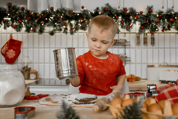 Cute little boy is very busy sifting flour through a sieve on the dough in Christmas decorated kitchen at home