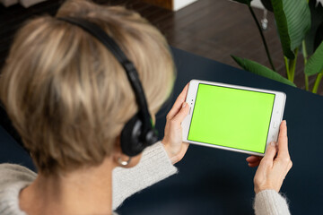 A middle-aged woman with a headset is viewing content using a tablet on a green screen, calling her relatives