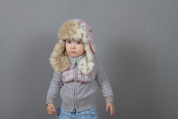 Cute baby girl 2 years old caucasian in warm winter clothes and a hat with earflaps with fur on a gray background