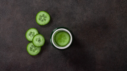 Cosmetic cream in a green jar with a vegetable cucumber on a black stone background