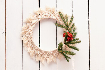 Creative Christmas wreath in the macrame style with fir branches and rowan berries is hanging on a white wooden wall,handmade decor in eco-style.Christmas, New Year and eco-friendly concept. - obrazy, fototapety, plakaty