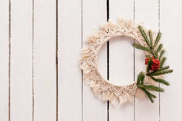 Creative Christmas wreath in the macrame style with fir branches and rowan berries is hanging on a white wooden wall,handmade decor in eco-style.Christmas, New Year and eco-friendly concept,copy space - obrazy, fototapety, plakaty
