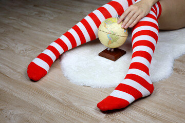 Female legs in Christmas knee socks, defocused view to woman sitting with globe on white fur rug. Concept of choosing travel tour for New Year celebration