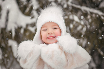 Childrens winter portrait, smiling happy girl in winter snowy forest in white fur coat and white hat