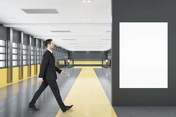 Businessman walking in modern black and orange tile shopping mall or airport interior with empty white mock up poster on wall, windows, city view and escalator. Public area concept.
