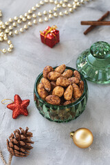 Candied or sugared almonds with honey and cinnamon in trendy green small glass jar on gray with Christmas decoration background. Traditional Christmas sweets for holidays. Copy space