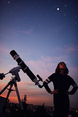 Girl with astronomical telescope stargazing under twilight sky.