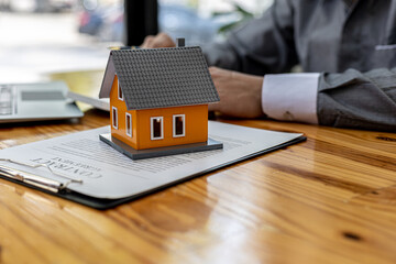 Replica of a small orange house sits on a table, a housing project salesperson is drafting a sales...