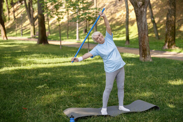 Cheerful senior woman does side bend holding plastic bar on in green park