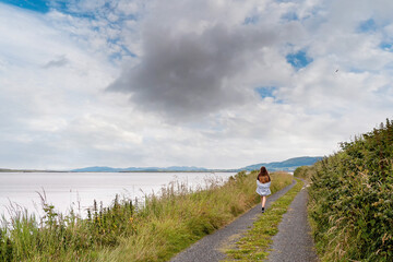 Teenager girl walking on a small country road with amazing view on the ocean and mountain and cloudy sky. County Sligo, Ireland. Knocknarea hill in the background. Travel and tourism concept