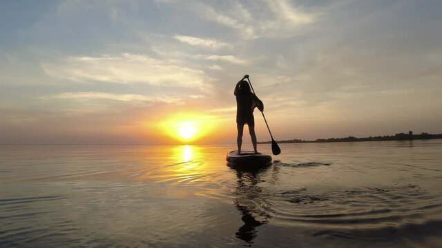 Female figure standing on a sup board and sailing into the setting summer sun.