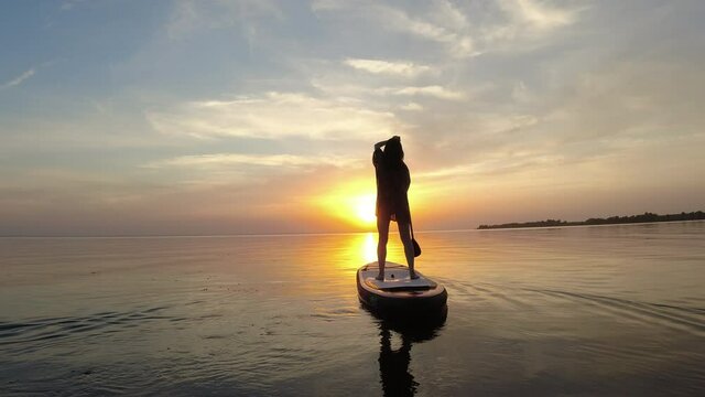 Female figure standing on a sup board and sailing into the setting summer sun.