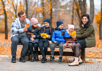 Fototapeta premium portrait of a family with children in an autumn city park - happy people sitting together on a wooden bench, posing against a background of beautiful yellow leaves