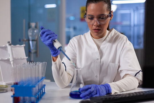 Specialist researcher dropping medical blue solution in glassware using micropipette working at biochemistry experiment in hospital laboratory. Doctor woman analyzing medicine results