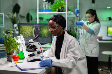 Naklejka na ściany i meble African american chemist woman writing medicine experiment results on clipboard after analyzing genetic mutation on plant sample under microscope. Scientist working in biochemistry laboratory