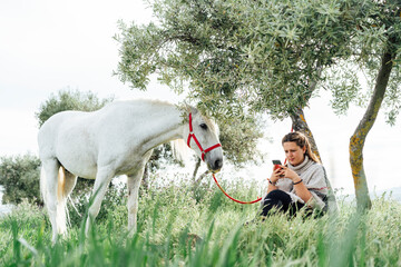Young woman using smart phone while sitting under tree by horse at field