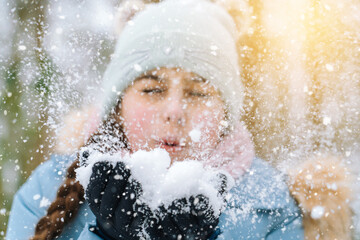 Girl playing on snow in winter time. Happy children in beautiful snowy winter forest on Christmas day. girl blows snow on snowflakes bokeh background. Happy childhood, active winter holidays concept 