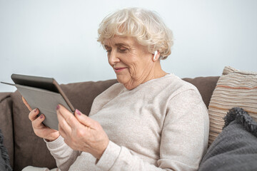 Grandmother wearing wireless earphones sitting on couch in living room and using tablet