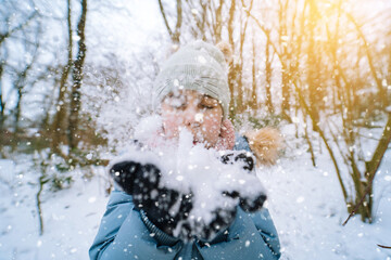 Fototapeta premium Girl playing on snow in winter time. Happy children in beautiful snowy winter forest on Christmas day. girl blows snow on snowflakes bokeh background. Happy childhood, active winter holidays concept 