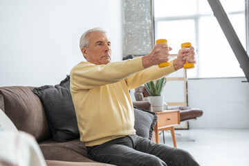 Old man doing exercise with dumbbells while sitting at the sofa and feeling serious