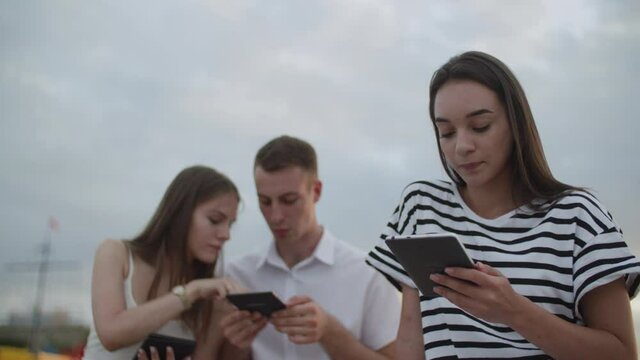 Two girls and a guy sitting in the park during the day reading an e-book