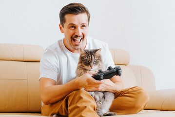 Happy young man having fun with angry cat playing video game at home. Cheerful guy player holding wireless joystick gamepad while sitting with pet on couch, indoors. Hobby, entertainment