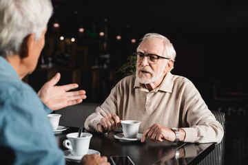 blurred man pointing with hand while talking to senior friend in cafe
