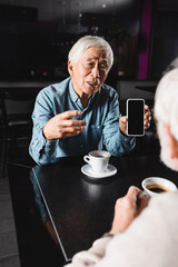 senior asian man pointing at cellphone with blank screen while spending time with blurred friend in cafe