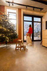 Woman with dog looking together on a snowy garden, standing back at cozy living room with Christmas tree. Concept of home comfort in winter. Idea of loneliness and celebration alone