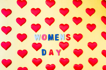 Multi-colored inscription Women's Day in the middle of rhythmically arranged silk red hearts on a yellow background. Postcard template for International Women's Day