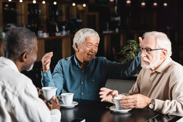 cheerful asian man gesturing during conversation with multiethnic friends in cafe