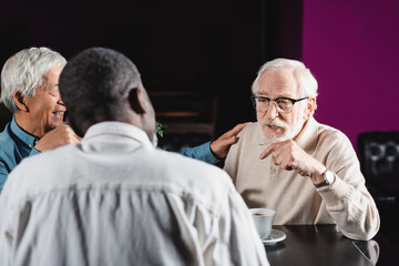 senior man in eyeglasses pointing with finger during conversation with interracial friends in cafe
