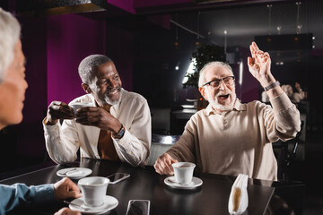amazed senior man looking away and waving hand while spending time with multiethnic friends in cafe