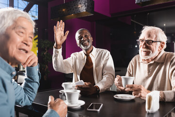 cheerful african american man looking away and waving hand near senior multiethnic friends in cafe