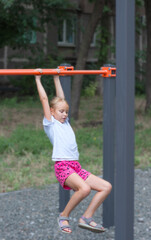 Obraz premium little girl 7 years old blonde hangs on a horizontal bar on a playground in a sleeping area