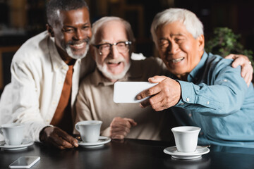 joyful multiethnic friends taking selfie on mobile phone in cafe, blurred background