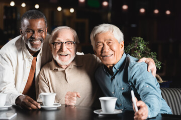senior multicultural friends embracing and smiling at camera near coffee cups in restaurant