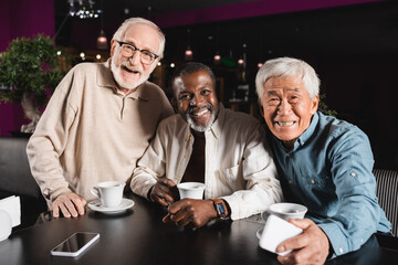 senior and cheerful multicultural friends looking at camera during meeting in restaurant