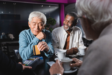 senior asian man paying near credit card reader in cafe near multiethnic friends