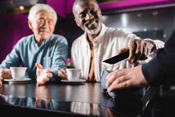 blurred senior man paying through terminal with mobile phone near asian friend