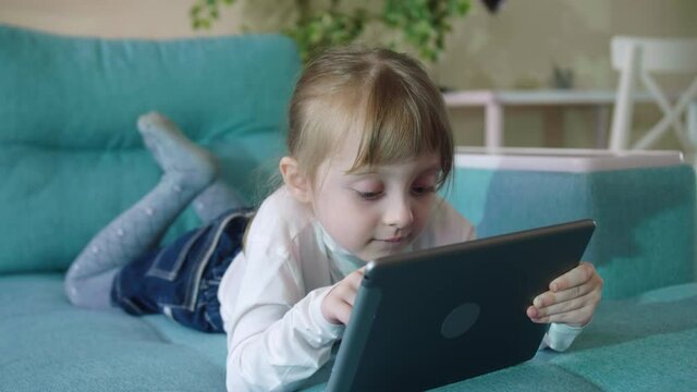 Preschooler Girl Playing Games Using Digital Tablet Lying On Sofa Alone. Curious Little Kid Girl Hold Pad Computer Watching Video At Home Children Education Or Tech Internet Addiction Concept
