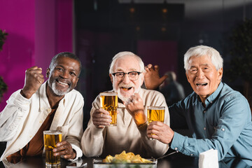 joyful senior multicultural friends with glasses of beer showing win gesture while watching football game in pub