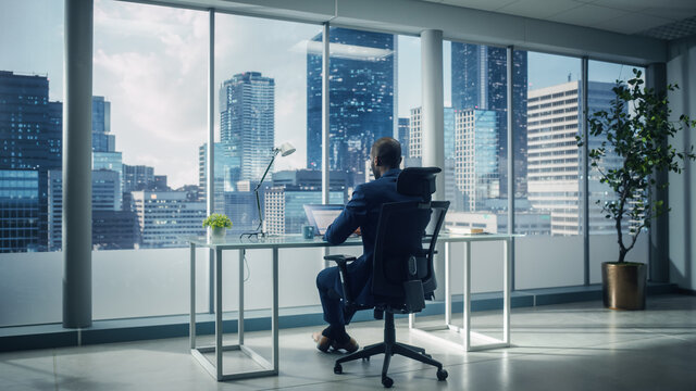 Successful African-American Businessman Sitting at His Desk Working on Laptop Computer in His Big City Office. Handsome Corporation CEO Planning Investment Strategy for e-Commerce Project.