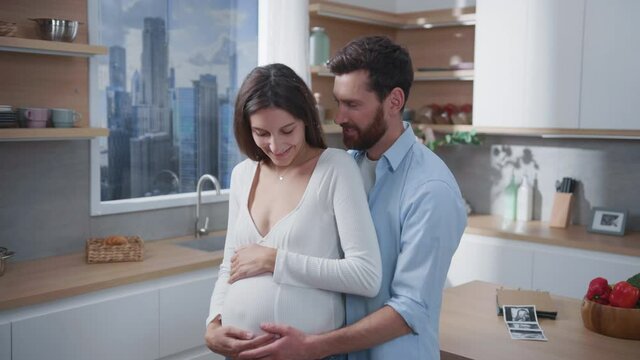 Affectionate couple of cheerful young woman pregnant with child hugging her husband enjoying each other staying home. Lovely kitchen. Loving family scene.