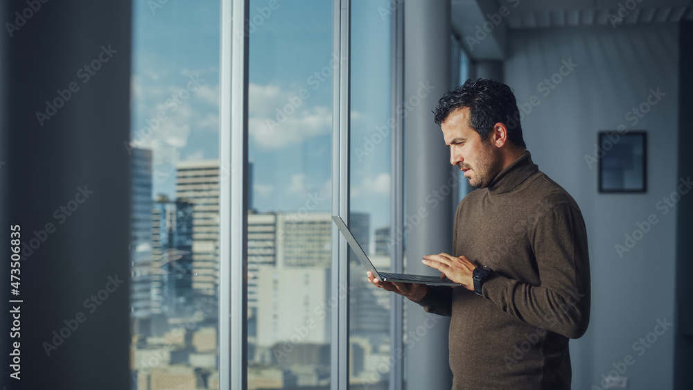 Poster thoughtful middle eastern businessman using laptop while standing in office near window on big city.