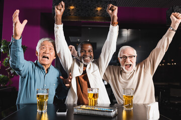 excited senior multiethnic friends watching football match in pub and showing win gesture