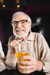 worried senior man in eyeglasses holding blurred glass of beer while watching soccer championship in pub